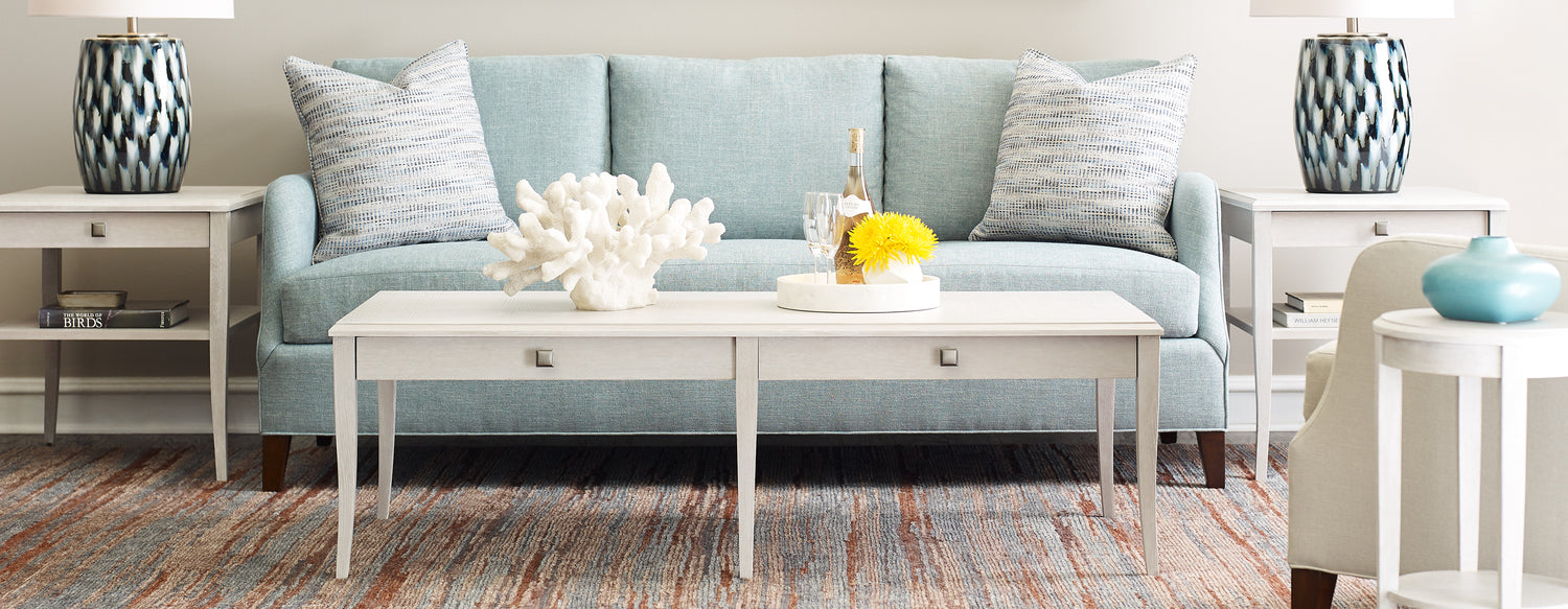 Origins by Stickley Harper light blue sofa with a gray pillow on each end, a white coffee table in front, and two white end tables on either side.