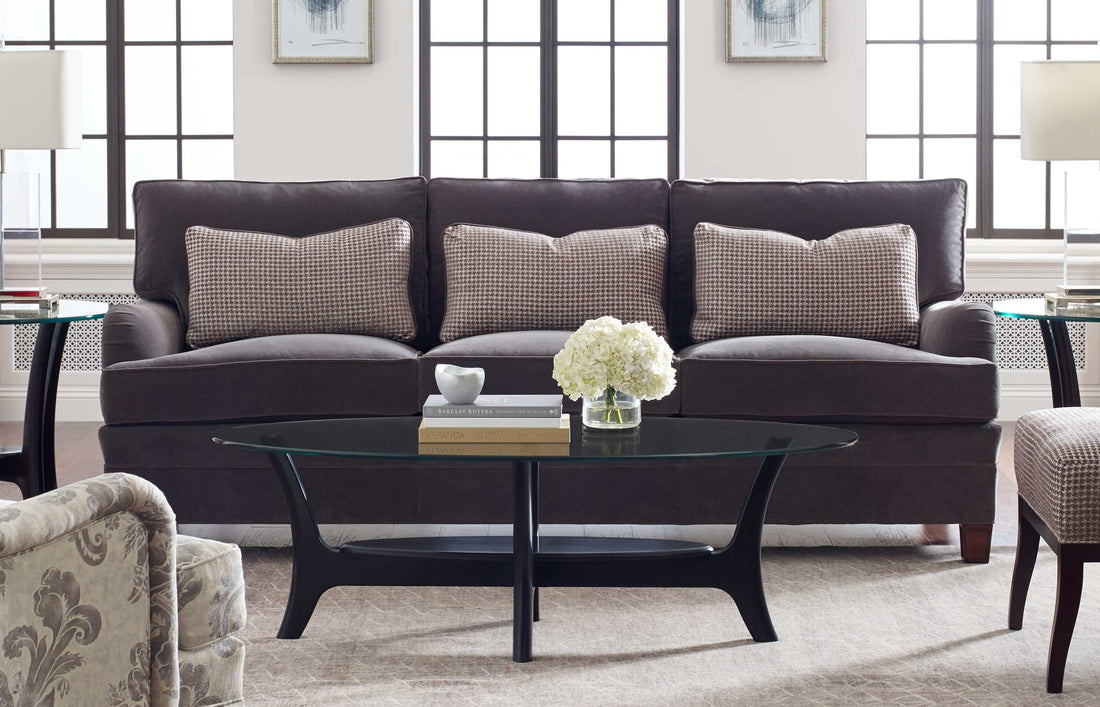 Lifestyle of a dark brown leather 7000 Series Three-Cushion Sofa with three accent pillows on it, there is a black glass-top cocktail table in front of it that has a small vase of white flowers on top