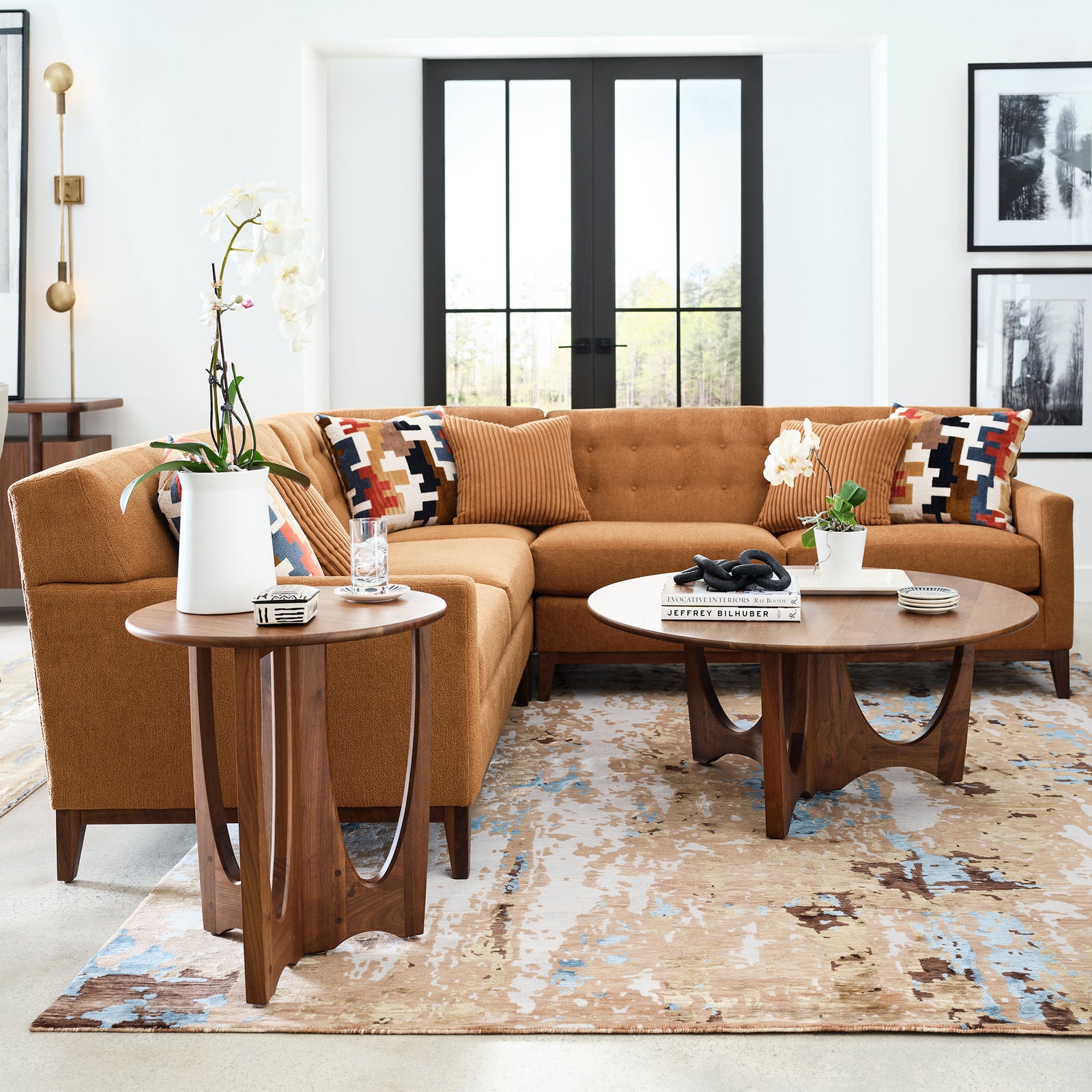 Walnut Grove living room featuring a mid-century modern sectional sofa in warm terracotta upholstery with tufted back cushions and geometric accent pillows, paired with a round walnut cocktail table and matching side table, both with sculptural cutout bases, styled on a textured area rug in a bright room with black French doors and framed wall art.