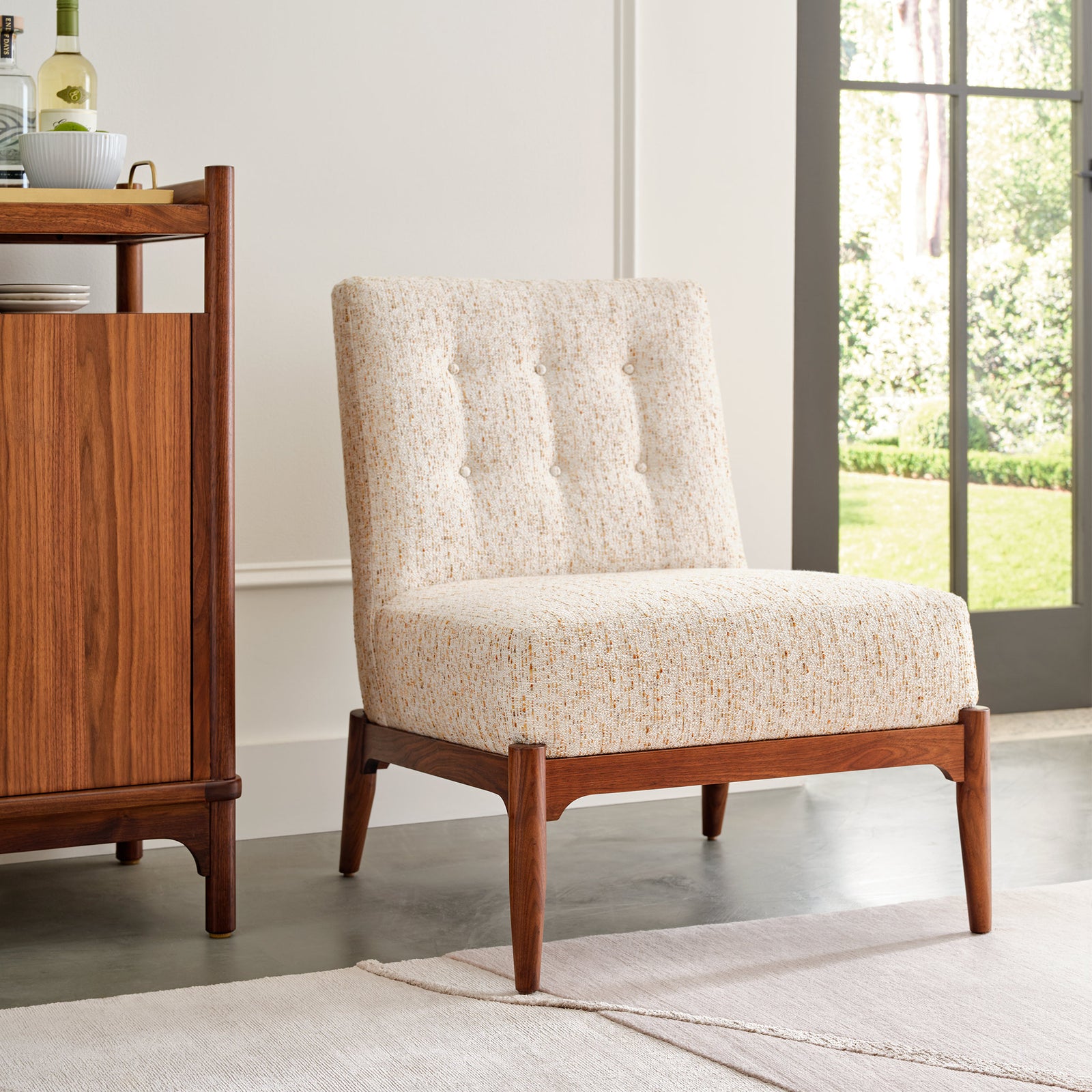 Stickley Walnut Grove Slipper Chair with a natural walnut base and tapered legs, upholstered in a textured cream fabric with subtle multicolor flecks and tufted button detailing, shown in a bright modern room with concrete floors and large windows overlooking a garden.