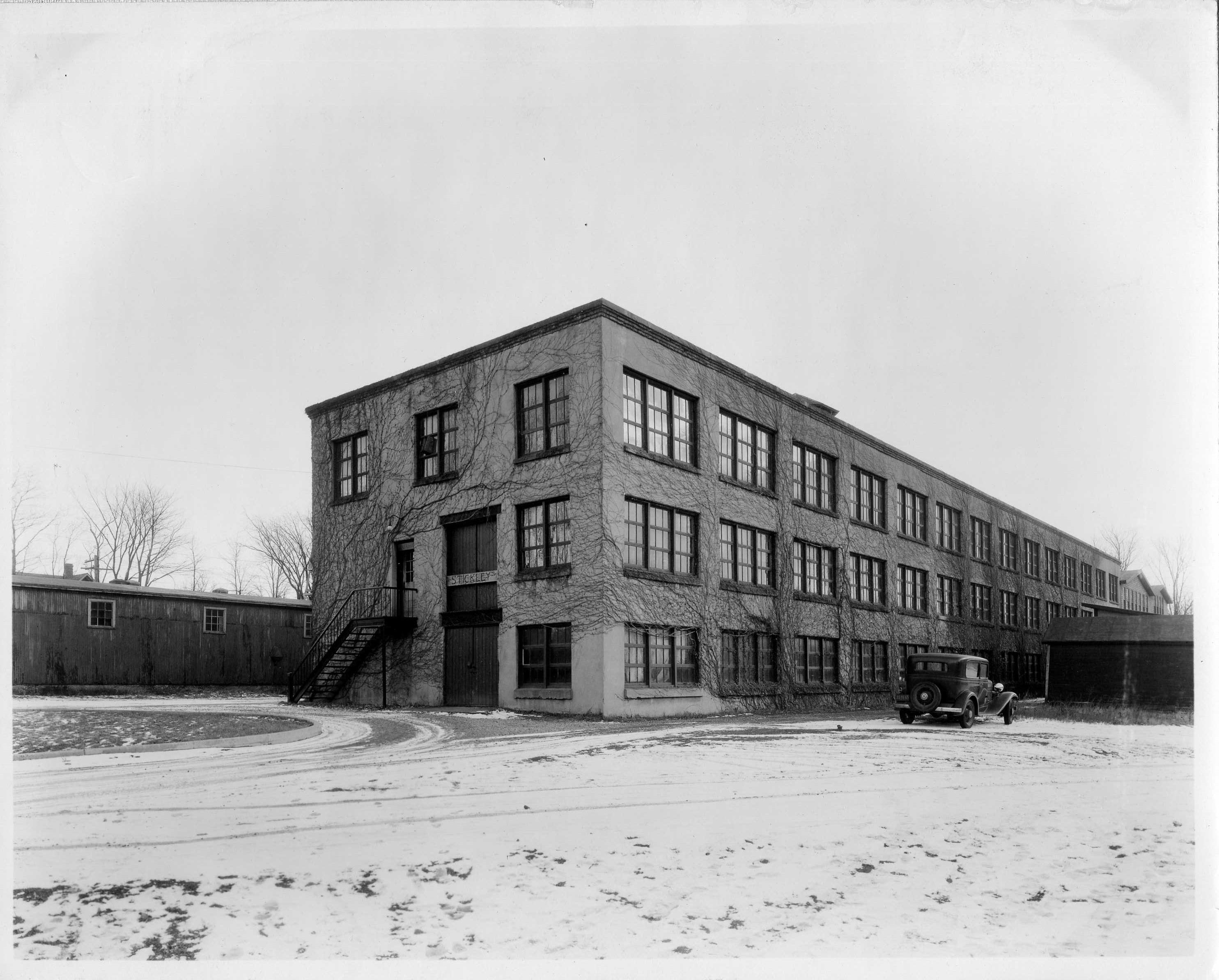 Exterior view of the original Stickley furniture factory in Fayetteville, New York, circa early 20th century, featuring ivy-covered brick walls, industrial windows, and a vintage car in a snowy setting.