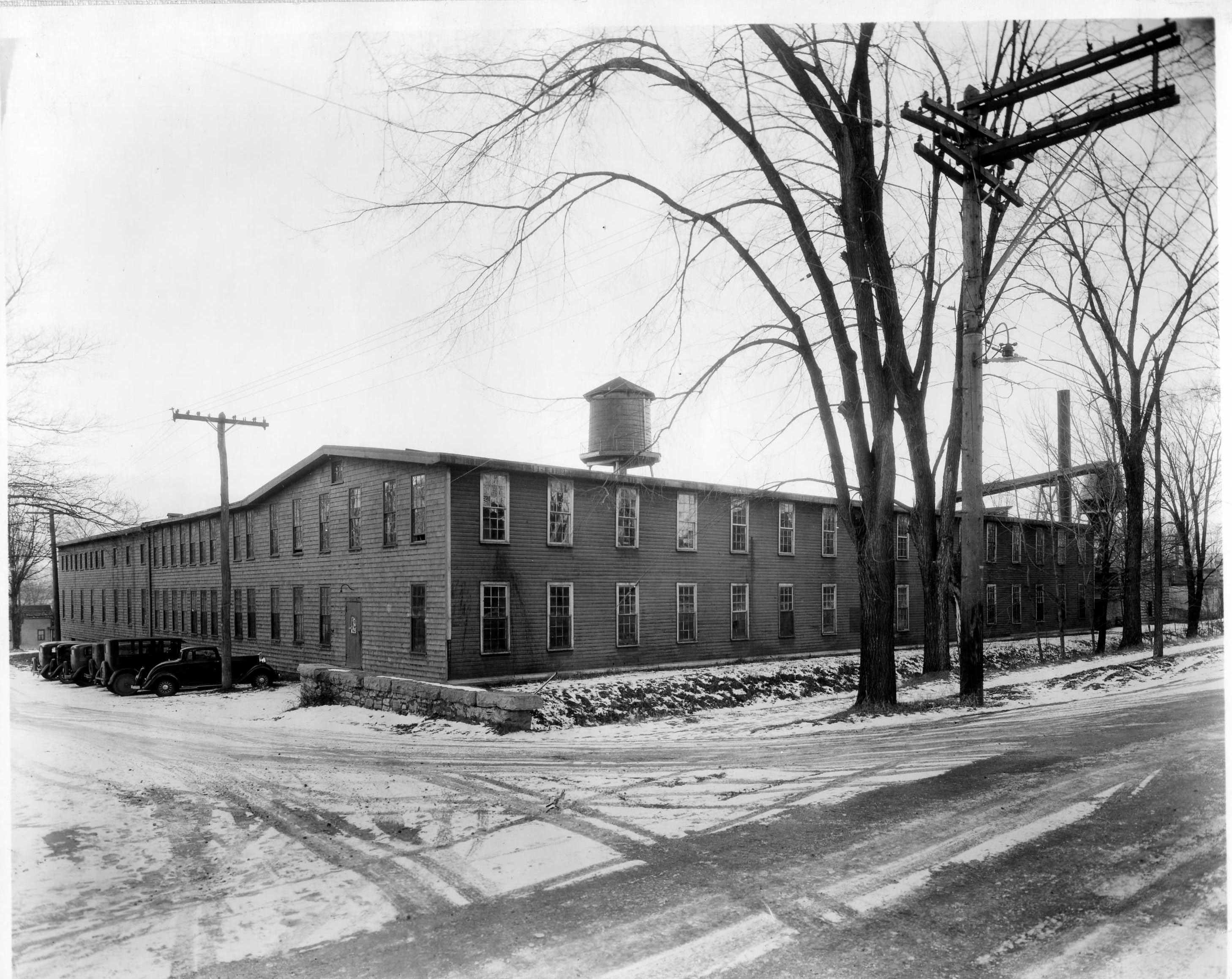 Archival photo of the Stickley furniture manufacturing plant in Fayetteville, NY, showing a two-story timber-frame factory with tall windows, vintage cars, a rooftop water tower, and snow-covered ground.