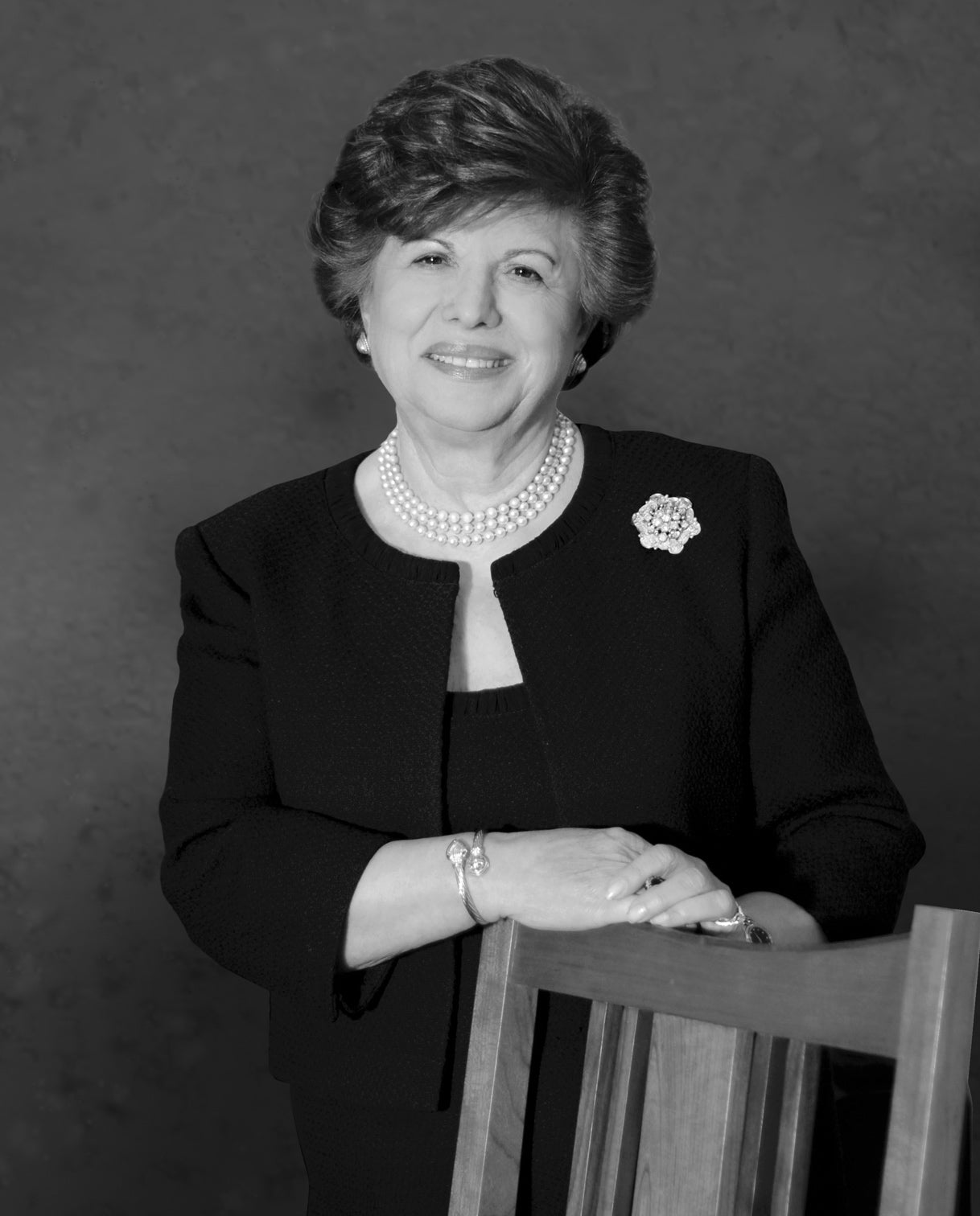 Black and white portrait of Aminy Audi standing beside a wooden chair, smiling gently. She is dressed in a dark suit with a pearl necklace and a floral brooch, exuding elegance and poise.