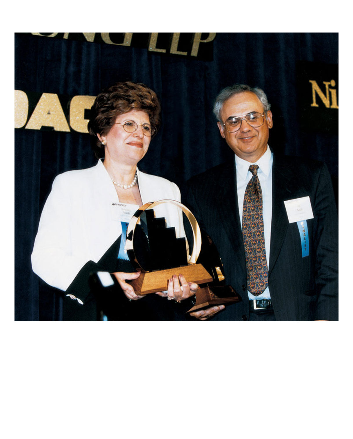 Aminy and Alfred Audi pose together at an awards ceremony. Aminy holds a distinctive award trophy featuring a circular and stair-step design. She wears a white blazer and pearls, while Alfred is in a dark suit and patterned tie. Both wear name tags and stand in front of a dark blue curtain with gold event signage.