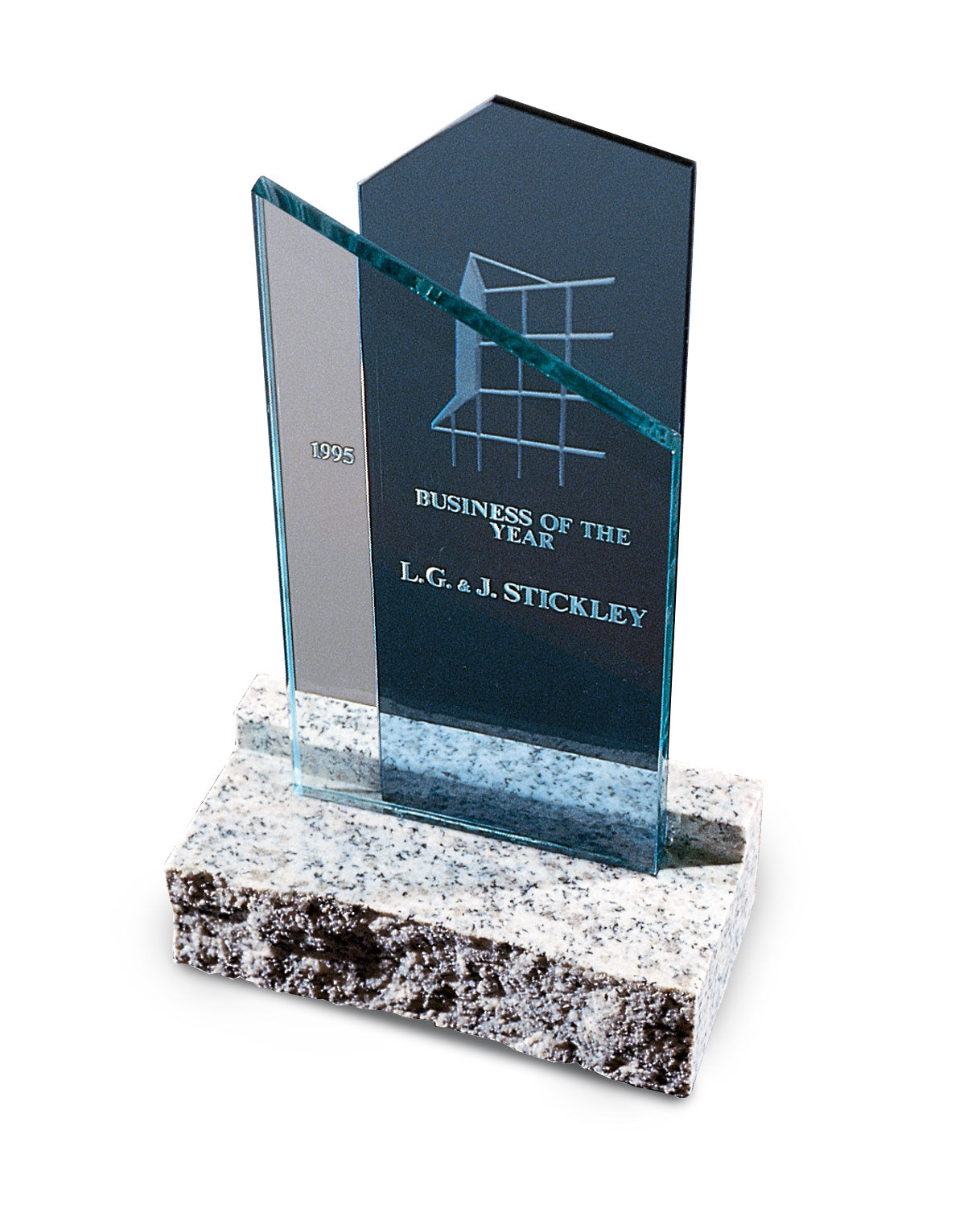 A clear glass award etched with the words "1995 Business of the Year – L. & J.G. Stickley" stands on a polished granite base. The award features a stylized grid design and a sleek, modern shape with two angled glass panels.