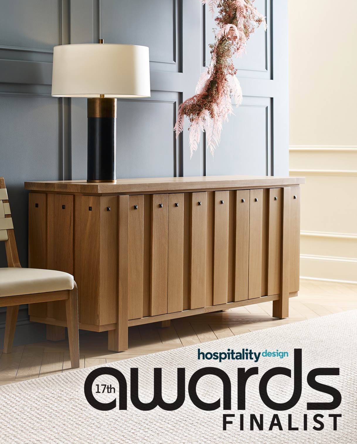 Hospitality Design 17th Awards Finalist image showcasing a light wood sideboard with vertical panel detailing and square metal hardware. The piece is styled in a sophisticated interior with blue paneled walls, a sculptural table lamp, and a delicate pink floral wreath overhead.