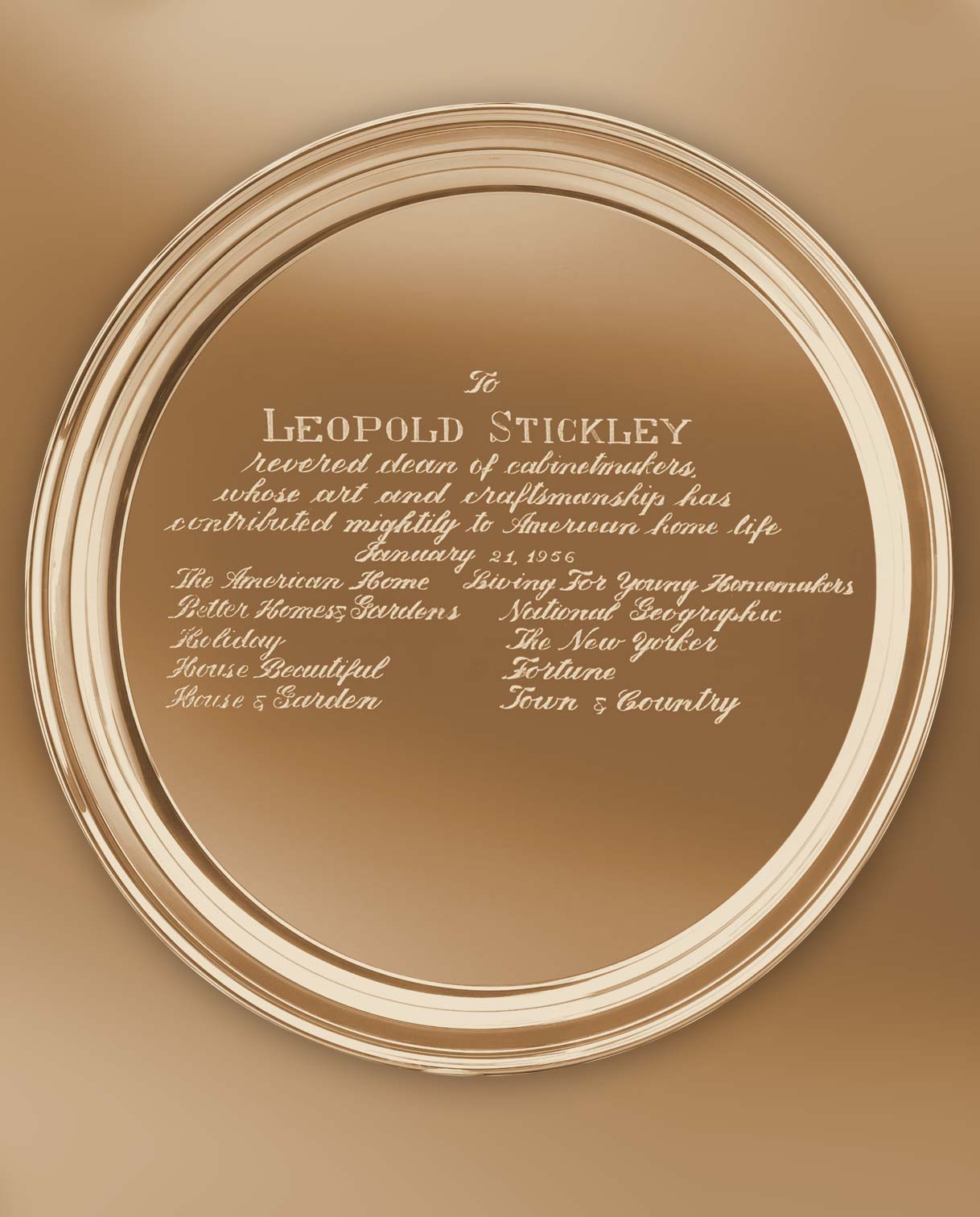 A commemorative gold-toned plaque awarded to Leopold Stickley on January 21, 1956. The inscription honors him as the "revered dean of cabinetmakers" whose craftsmanship significantly contributed to American home life. The plaque is signed by various notable publications including Better Homes & Gardens, National Geographic, The New Yorker, and House Beautiful.