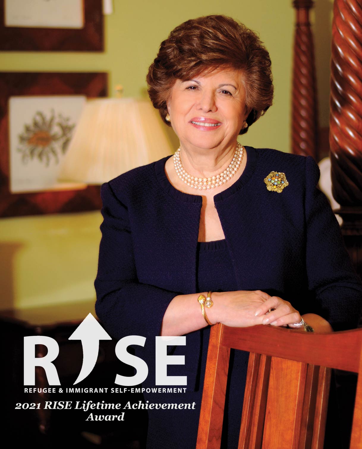 Portrait of Aminy Audi smiling and dressed in formal attire, wearing pearls and a navy blazer with a brooch, posed beside a wooden chair. The image features the RISE logo with the text “2021 RISE Lifetime Achievement Award,” recognizing her for her contributions to Refugee & Immigrant Self-Empowerment.