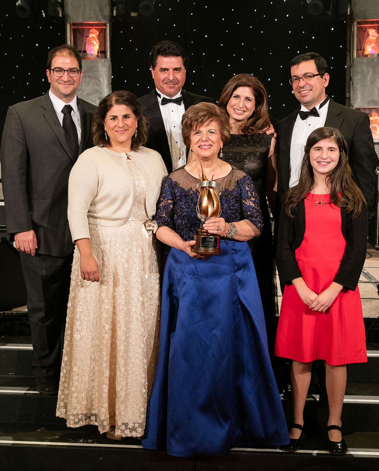 Aminy Audi poses with her family at an awards ceremony, holding a distinguished trophy. She wears a royal blue gown with a lace bodice, surrounded by family members dressed in formal evening attire, standing on a stage with a starry backdrop.