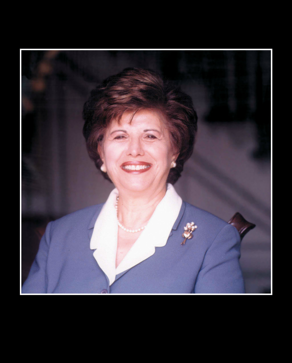 Portrait of Aminy Audi smiling warmly in a blue blazer with a white collar, adorned with a pearl necklace and a decorative brooch, seated in front of a softly lit background.