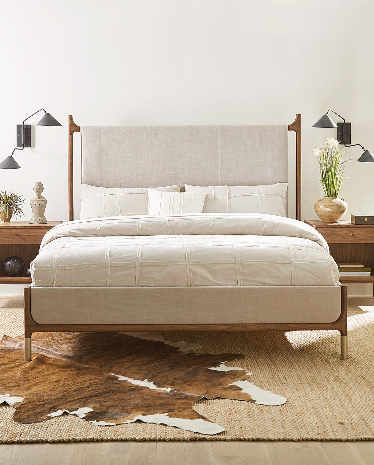 Modern minimalist bedroom showcasing the Walnut Grove bed with a sleek walnut frame and upholstered headboard and footboard in a soft neutral fabric. The bed is styled with crisp white bedding and flanked by matching walnut nightstands, each topped with sculptural decor, fresh greenery, and matte black wall-mounted reading sconces. A woven natural fiber rug layered with a cowhide accent rug adds warmth and texture to the serene, contemporary space.