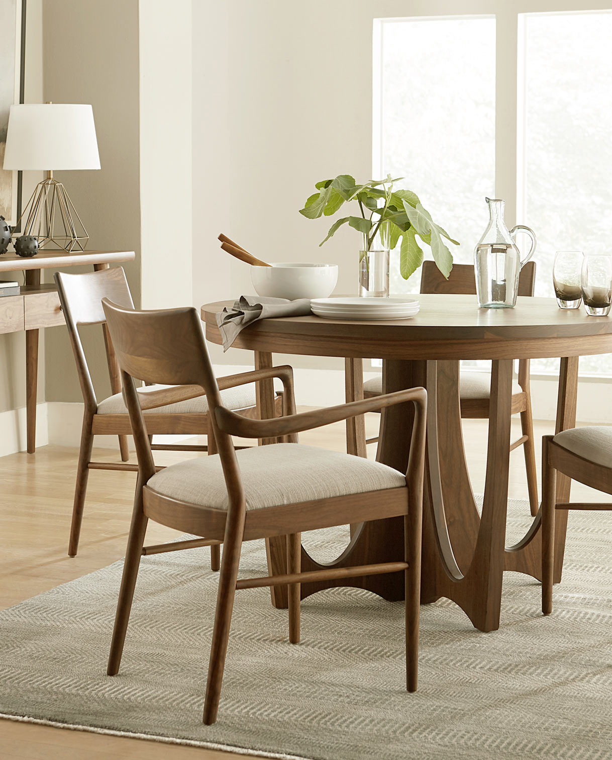 Walnut Grove dining set lifestyle featuring a round walnut pedestal table surrounded by sculpted wood chairs with upholstered seats. Set in a softly lit dining room with minimalist decor, neutral tones, and natural textures, the table is styled with a glass pitcher, plates, and a leafy centerpiece for a warm, modern aesthetic.