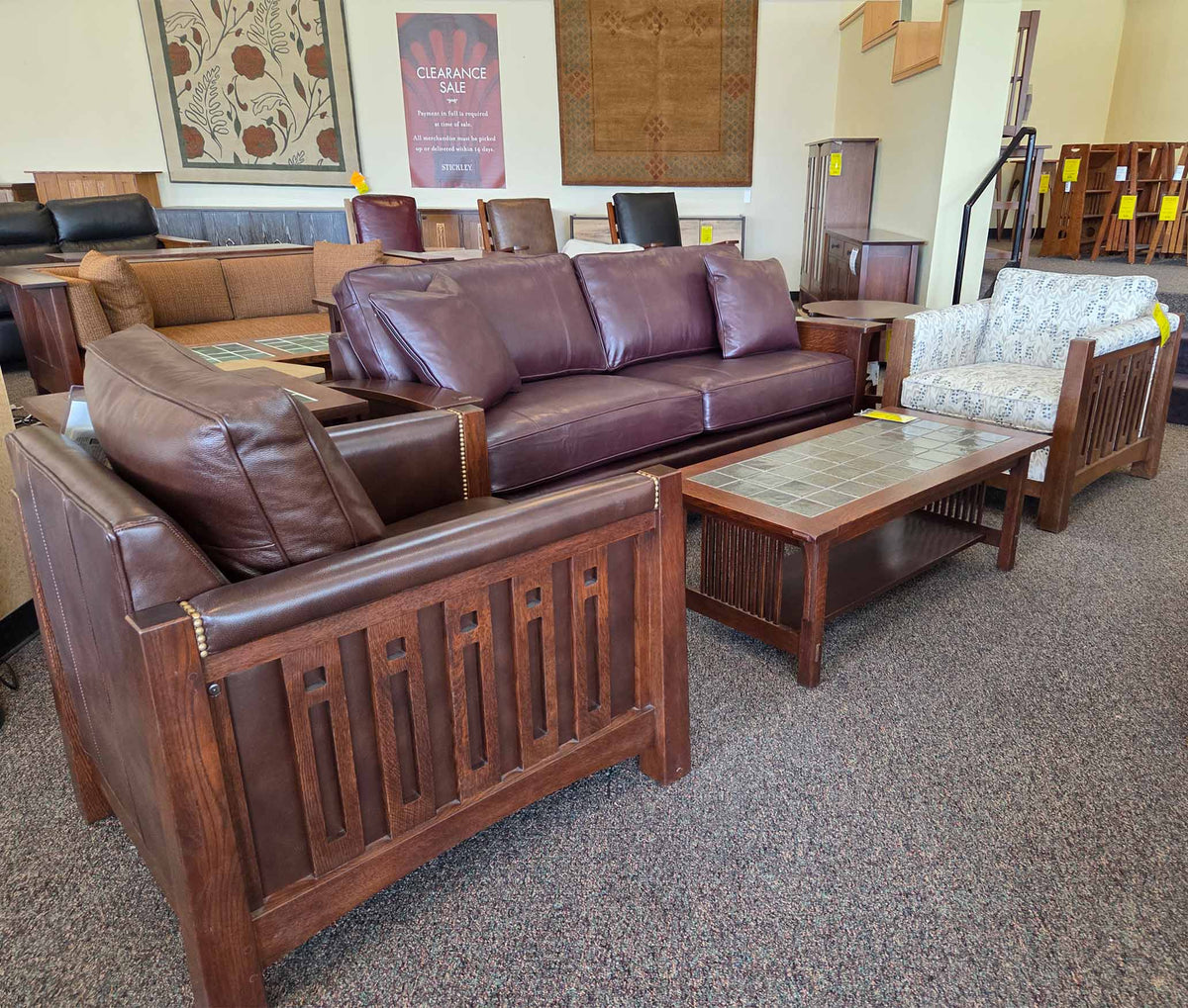 Interior of the North Syracuse Stickley Furniture Outlet showing a variety of Mission-style furniture, including a brown leather sofa and chair with slatted wood frames, a tile-top coffee table, and a patterned upholstered chair, all arranged in a carpeted showroom with clearance signage.