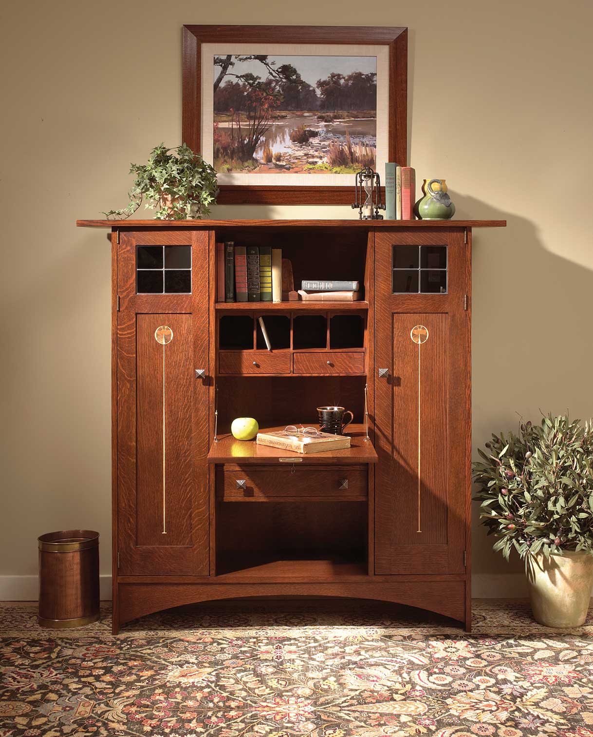 The Stickley Harvey Ellis Fall-Front Bookcase with Wood Doors, crafted in rich oak with intricate inlay and leaded glass cabinet fronts. Shown in a warm home office setting with books, accessories, and a framed landscape painting above, highlighting its functional drop-down writing surface and classic Arts and Crafts design.