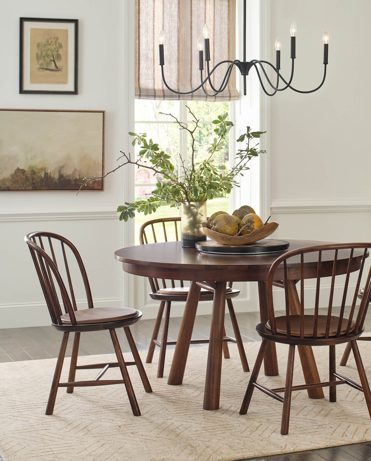 Hudson Valley dining room lifestyle showcasing a round natural walnut dining table with softly tapered legs, paired with matching spindle-back chairs. The room is styled with a modern black chandelier, botanical wall art, and a neutral rug for a warm, inviting setting. A centerpiece of leafy branches and wooden bowl adds an organic touch.