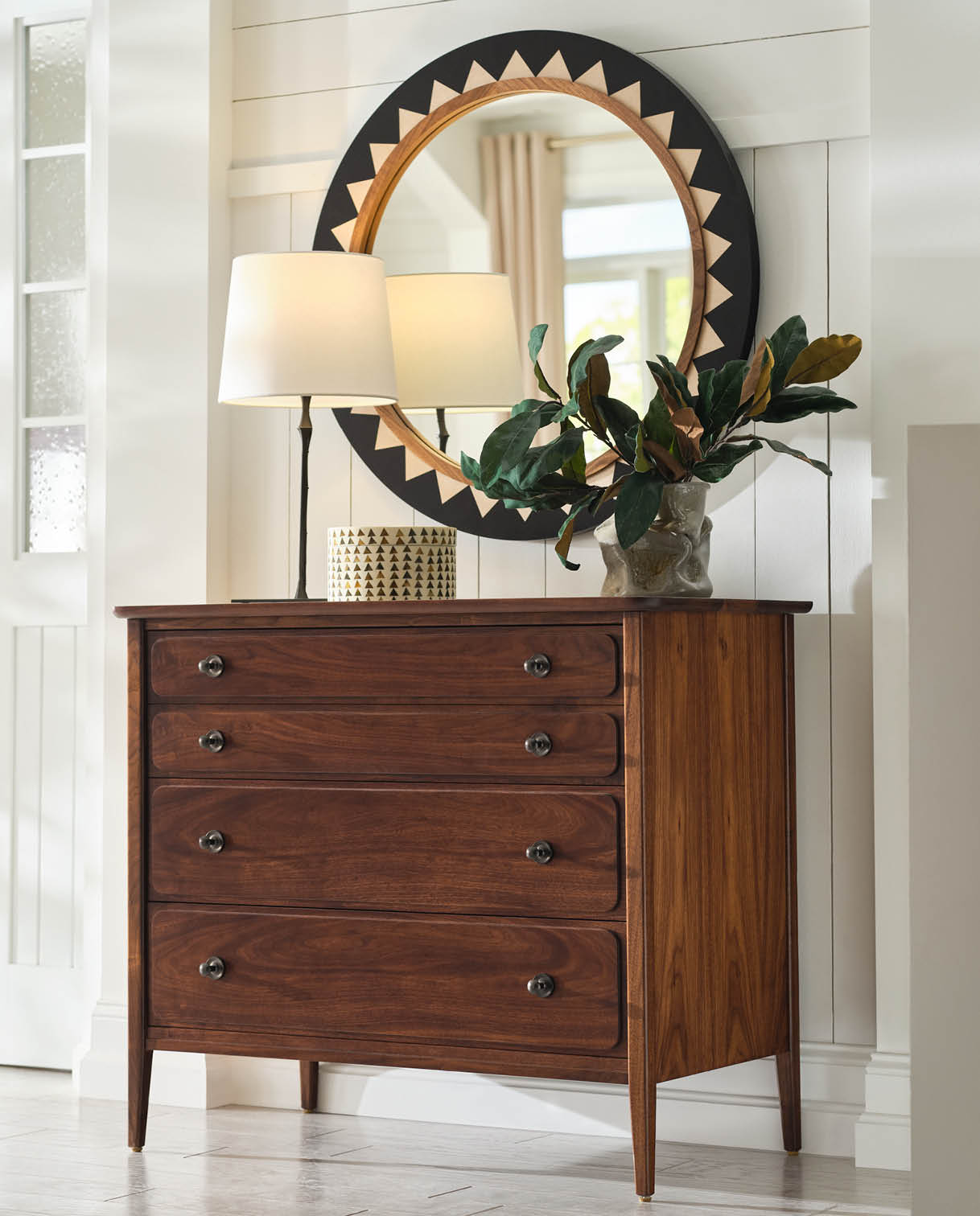 Hudson Valley four-drawer walnut dresser with rounded fronts and dark metal knobs, styled with a modern ceramic lamp, decorative vase of magnolia leaves, and a graphic round mirror, set in a bright bedroom with white shiplap walls and natural light.