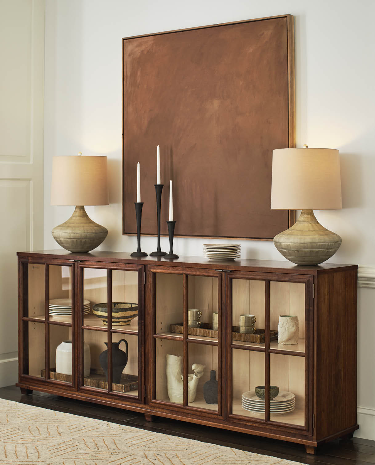 Hudson Valley glass-front dining server in rich walnut, featuring three cabinet sections with grid-style wood doors, styled with ceramic vases, dishware, and trays, and topped with sculptural lamps and black candleholders beneath a large bronze abstract painting.