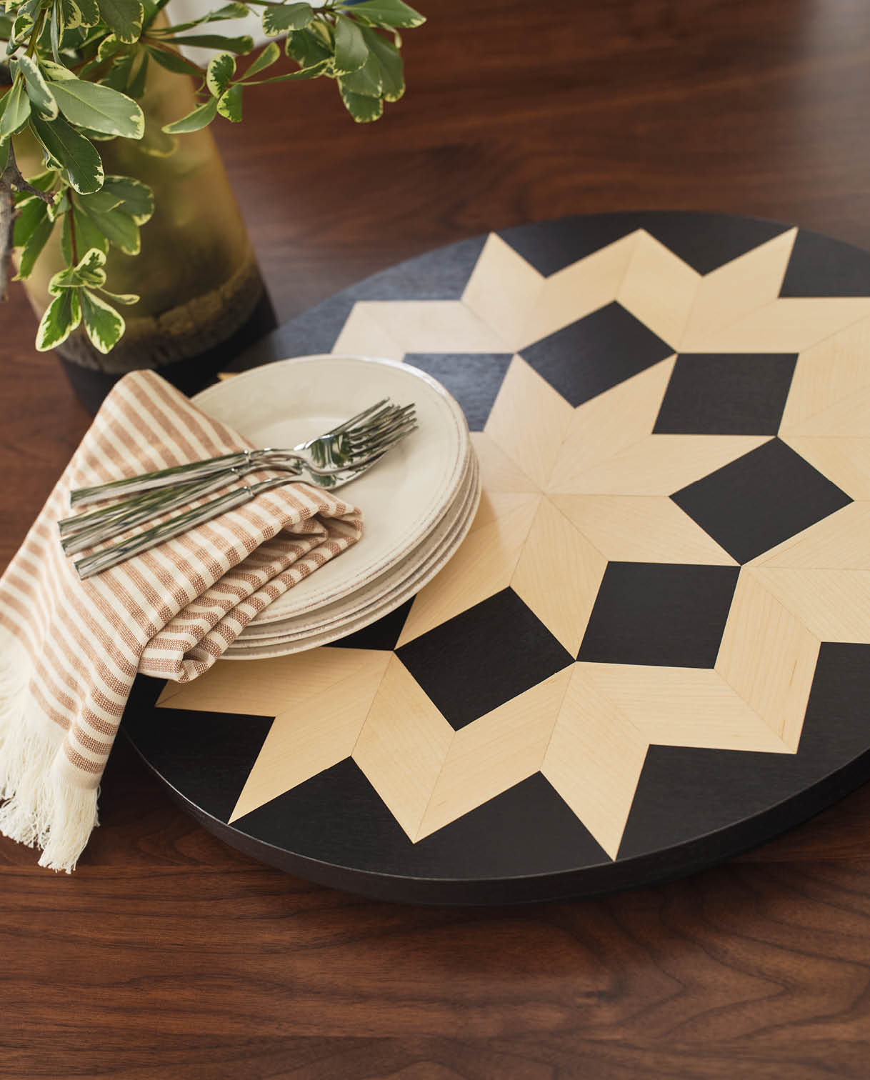 Stickley Hudson Valley Lazy Susan with a bold geometric starburst pattern in natural and black wood tones, styled on a walnut dining table with stacked ceramic plates, striped cloth napkin, silver forks, and a green glass vase with variegated foliage.