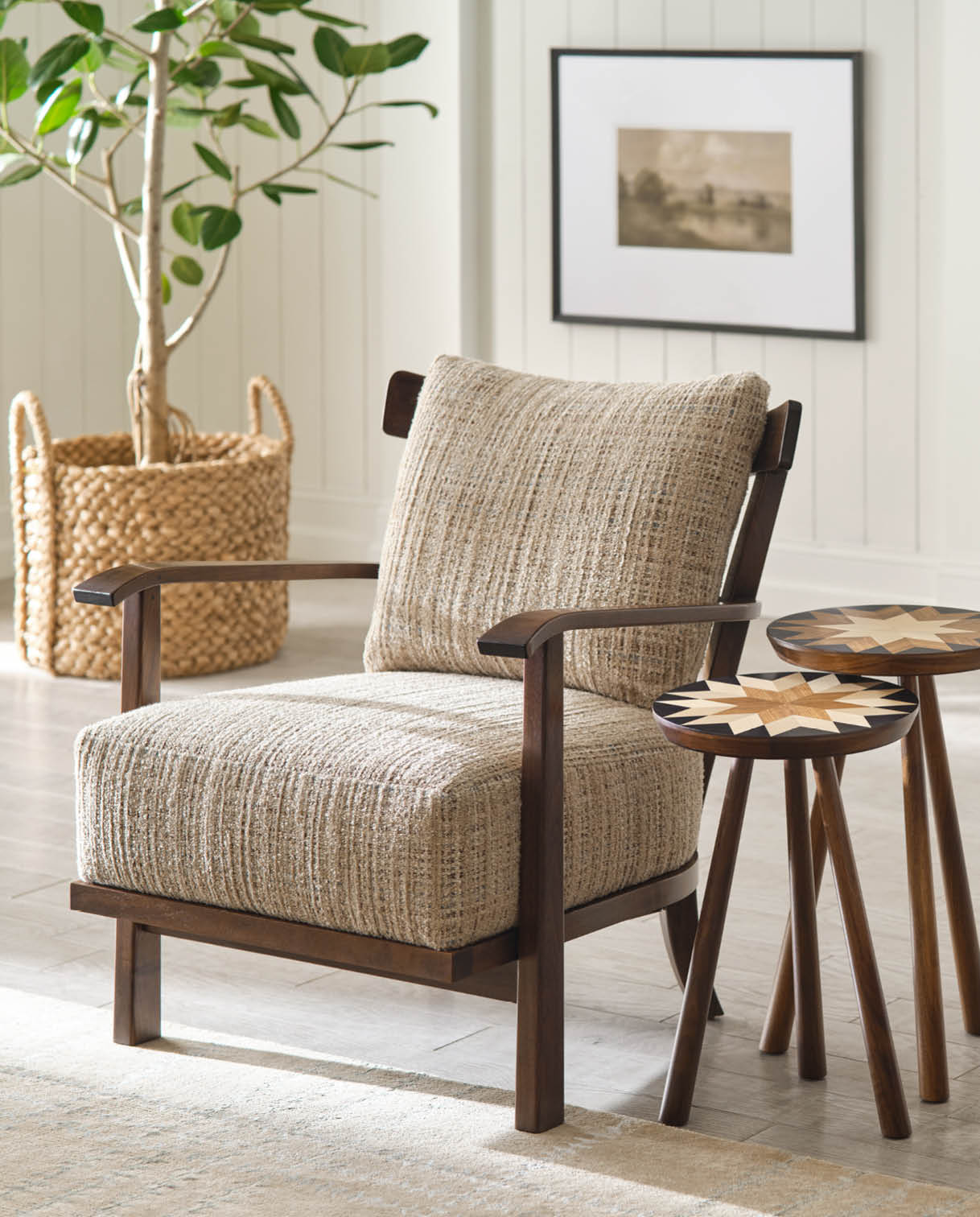 Stickley Hudson Valley accent chair with textured beige upholstery and a dark wood frame, shown next to two round starburst-inlay drink tables in a bright living room with light wood floors, shiplap walls, and a woven basket planter with a tall leafy plant.