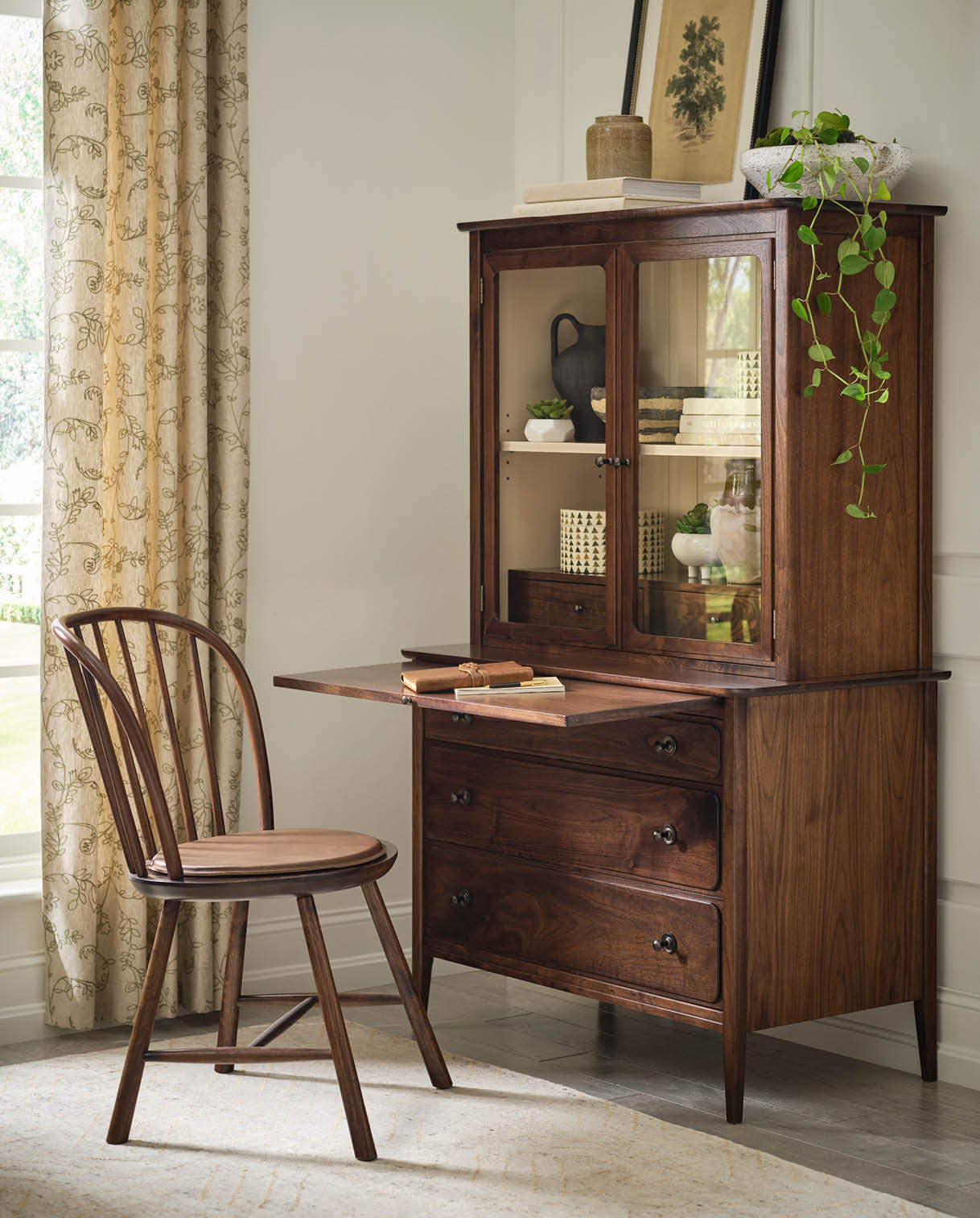 Stickley Hudson Valley secretary desk in warm walnut with a drop-front writing surface, three lower drawers, and an upper hutch with glass doors displaying books and ceramics, paired with a matching Windsor-style side chair in a bright home office with floral curtains and soft natural light.