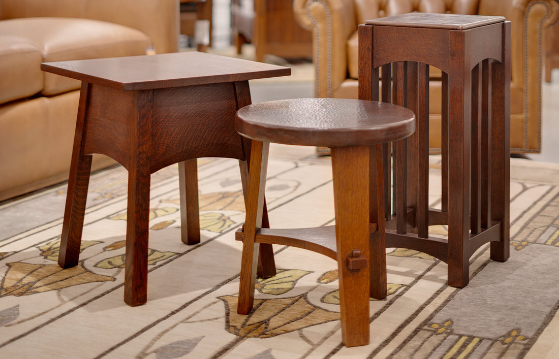 Three handcrafted Stickley Mission-style accent tables, including a rectangular table, a round copper-topped table, and a tall slatted pedestal table, displayed on an Arts & Crafts floral rug with leather furniture in the background.