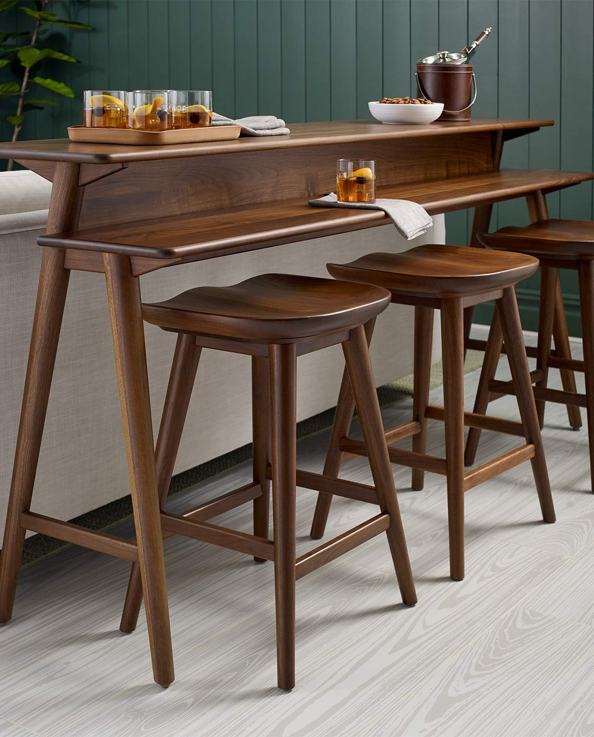 Modern home bar setup featuring a sleek walnut counter-height console table with sculpted edges and matching wood barstools. The setup is styled with cocktails, linen napkins, a bowl of snacks, and an ice bucket, creating a welcoming entertaining space. Set against a green paneled accent wall and light wood flooring, the design combines mid-century inspiration with contemporary elegance.