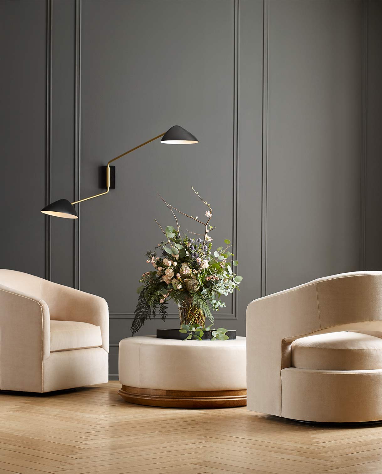 Lifestyle of two Portfolio120 Amelia Chairs with a Winona Round Ottoman between them with a large floral bouquet on top, they are against a dark gray wall