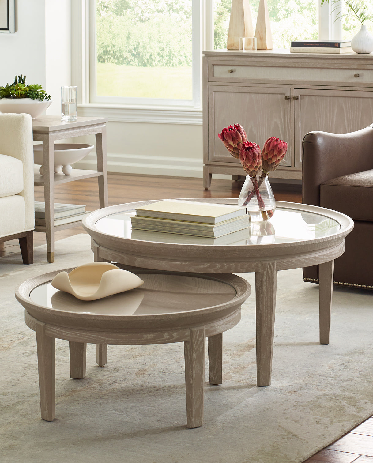 Pair of Maidstone Round Cocktail Tables by Stickley in a soft whitewashed oak finish, featuring round glass tops and tapered legs, styled in a light-filled living room with neutral furnishings and decorative accents for a modern coastal aesthetic.