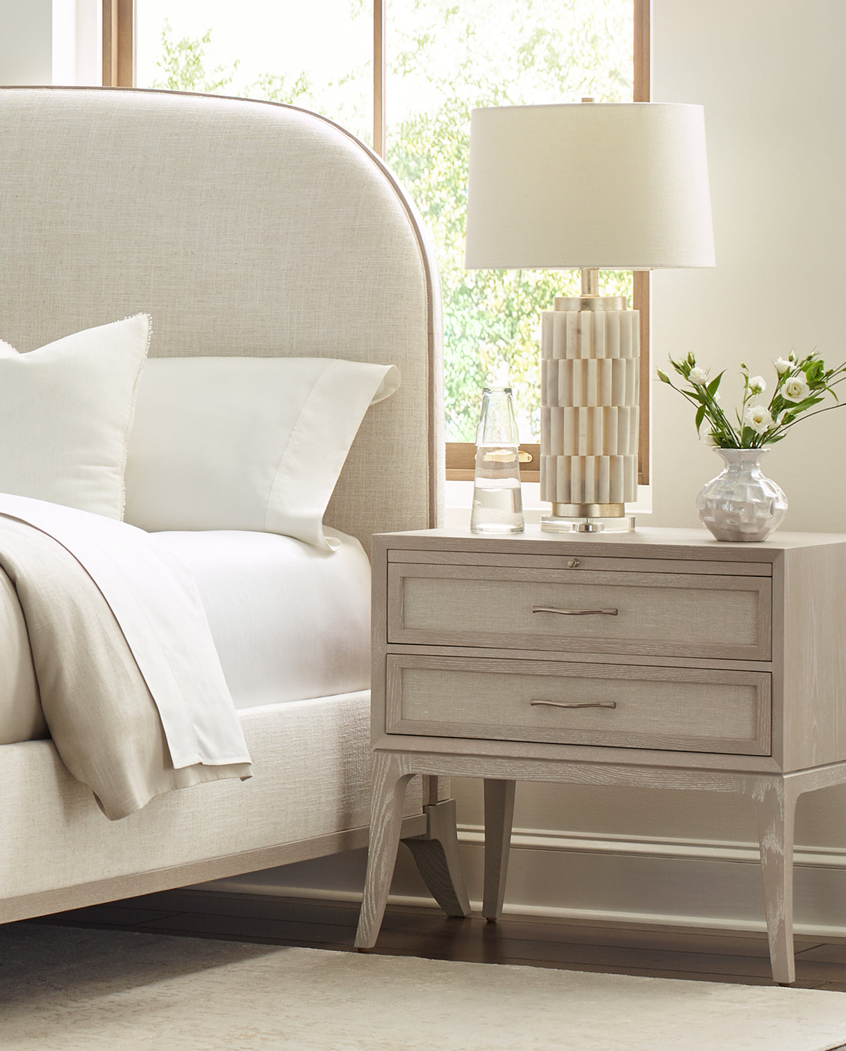 Stickley Maidstone bedroom featuring an upholstered bed with a softly curved headboard and textured linen fabric, paired with a two-drawer nightstand in a light oak finish with woven jute drawer fronts and elegant metal hardware.