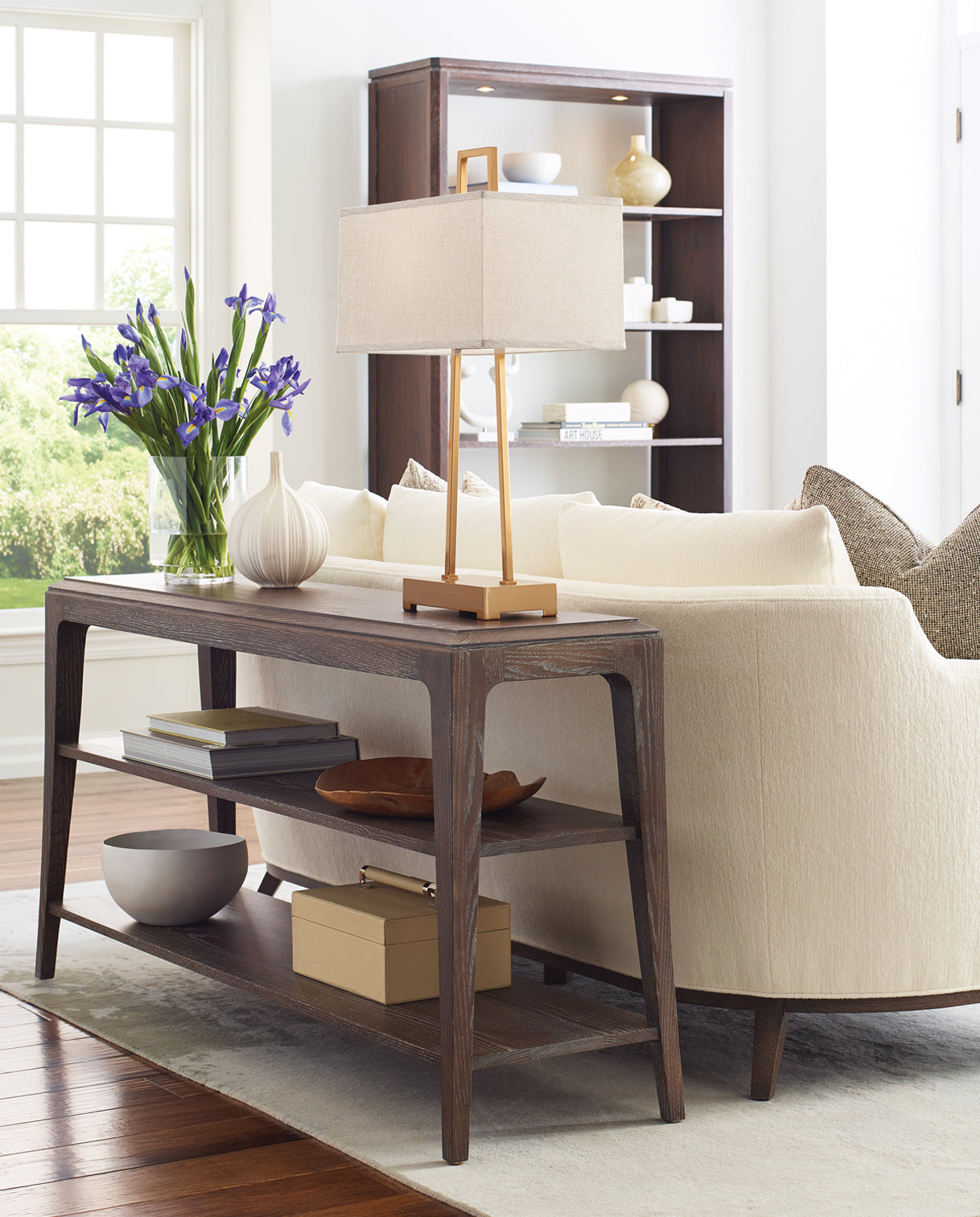 Stickley Maidstone living room featuring a sculptural wood sofa table in a soft charcoal finish with open shelving, styled behind a curved cream upholstered sofa with textured accent pillows, accompanied by a modern rectangular lamp, a vase of purple irises, and a minimalist display cabinet in the background.