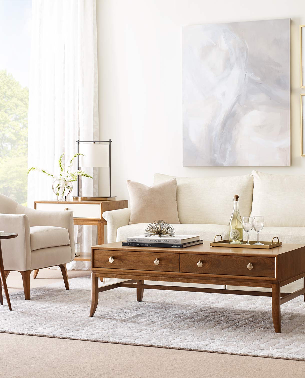 A Martine Cocktail table sits in front of a white upholstered couch and accent chair, there is a decorative tray with wine glasses on top of it