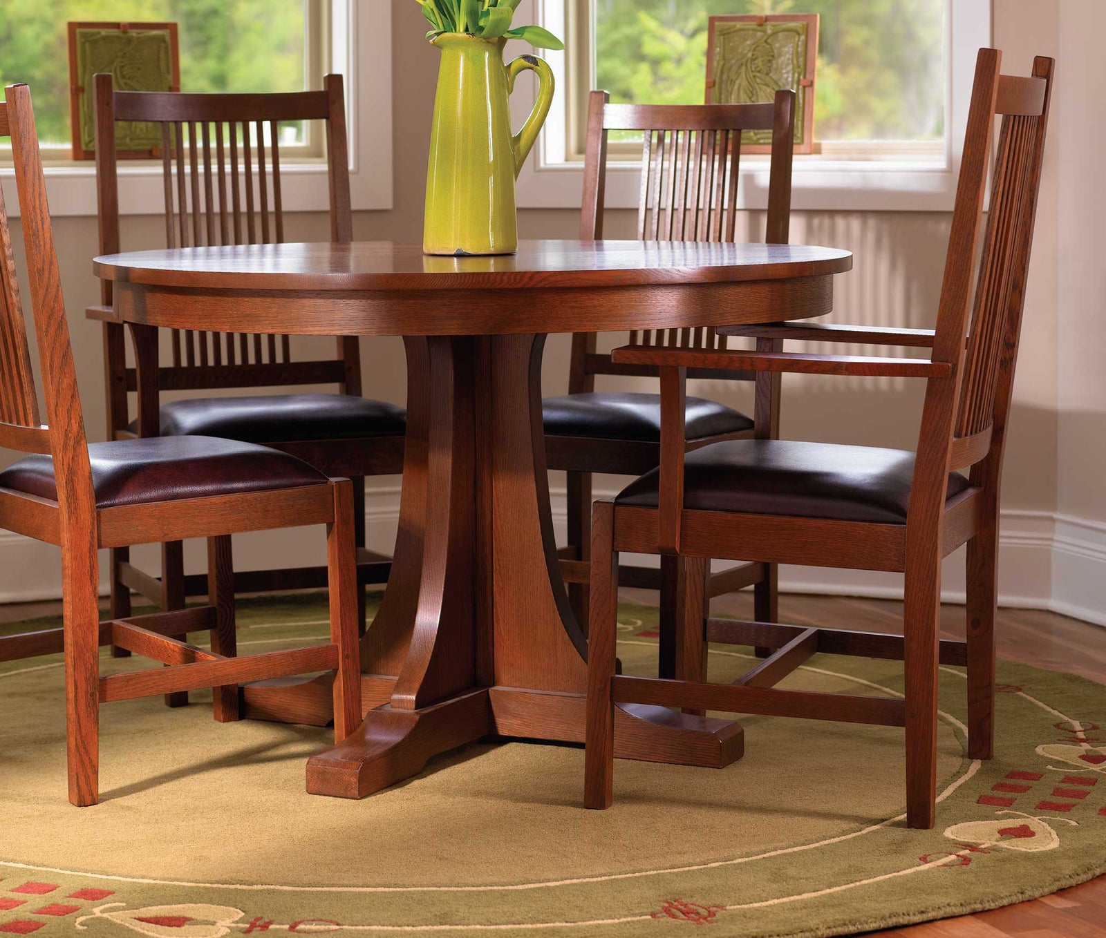 Stickley Mission Collection dining room featuring a round pedestal dining table and slatted-back dining chairs with dark leather seats. The furniture is crafted in rich oak wood with a warm finish, showcasing classic Arts and Crafts styling. A bright green pitcher with flowers adds a pop of color to the table, and the setting is grounded by a circular green rug with a decorative border. Natural light streams in through the surrounding windows, highlighting the timeless craftsmanship and elegance of the room