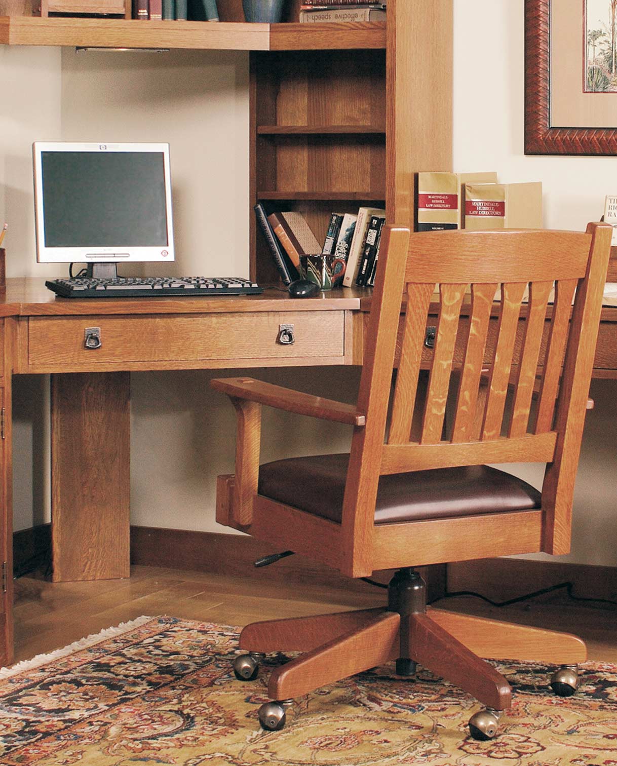 Stickley Mission collection home office setup featuring a solid oak desk and matching slat-back swivel desk chair with leather seat cushion, set on a traditional patterned rug with a built-in hutch and computer monitor.
