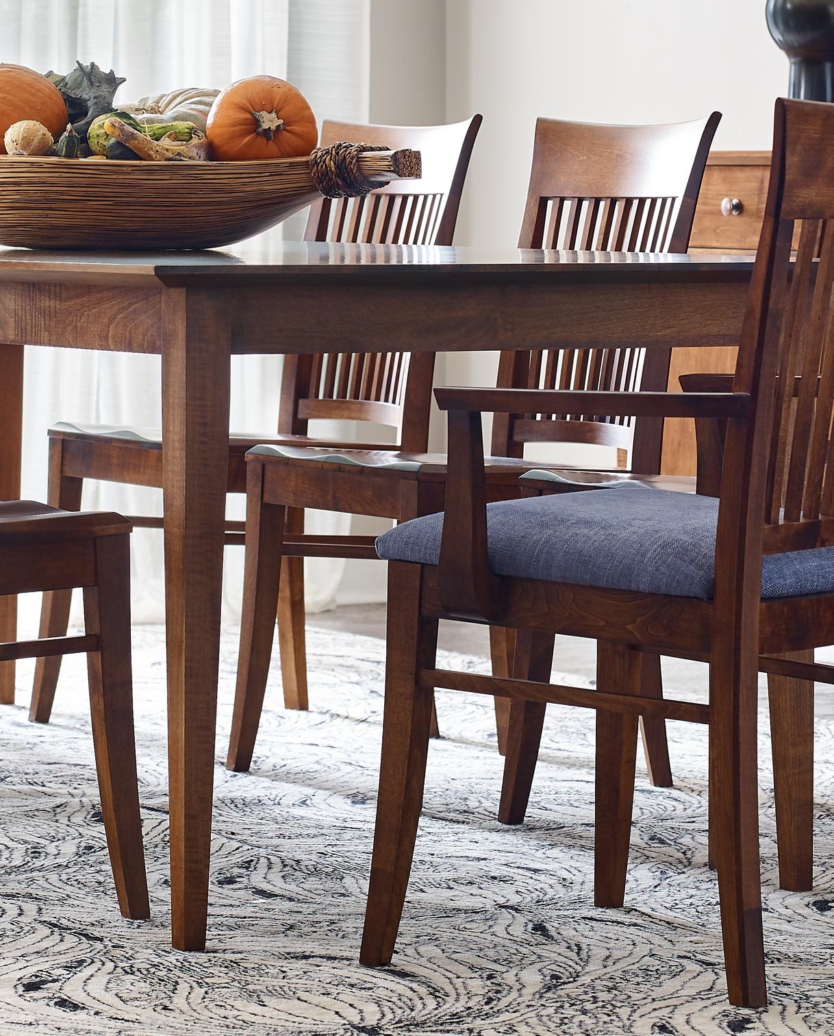 Lifestyle of a Gable Road Dining Table with a large basket full of gourds on top of it. Eight matching chairs surround the table and there is a Gable Road Large Server behind it against a white wall.