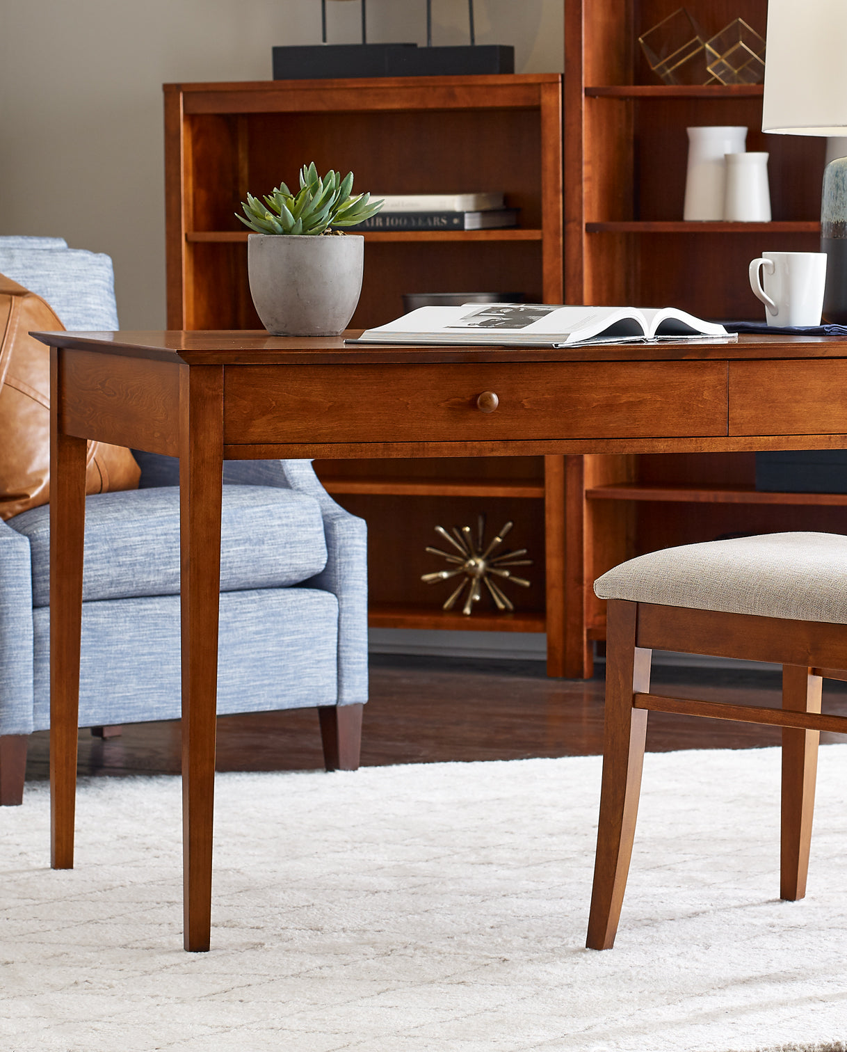 Lifestyle of a Gable Road Desk that has an opened book on top of it as well as a small potted plant and teacup, there is a matching Gable Road Chair pulled out from it