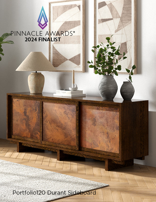 Stylish interior showcasing the Pinnacle Awards® 2024 Finalist, the Portfolio120 Durant Sideboard, featuring a rustic wood frame with sliding copper-toned panel doors. The sideboard is topped with a ceramic lamp, stacked books, and modern vases with greenery, and sits against a white wall with abstract framed art.