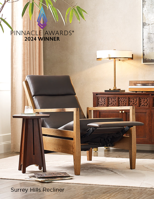 Elegant living space featuring the award-winning Surrey Hills Recliner with light wood frame and dark leather cushions, shown in a reclined position next to a sculptural wood side table with a glass of water; in the background is a carved wood sideboard with a modern table lamp. Text overlay reads “Pinnacle Awards® 2024 Winner.”