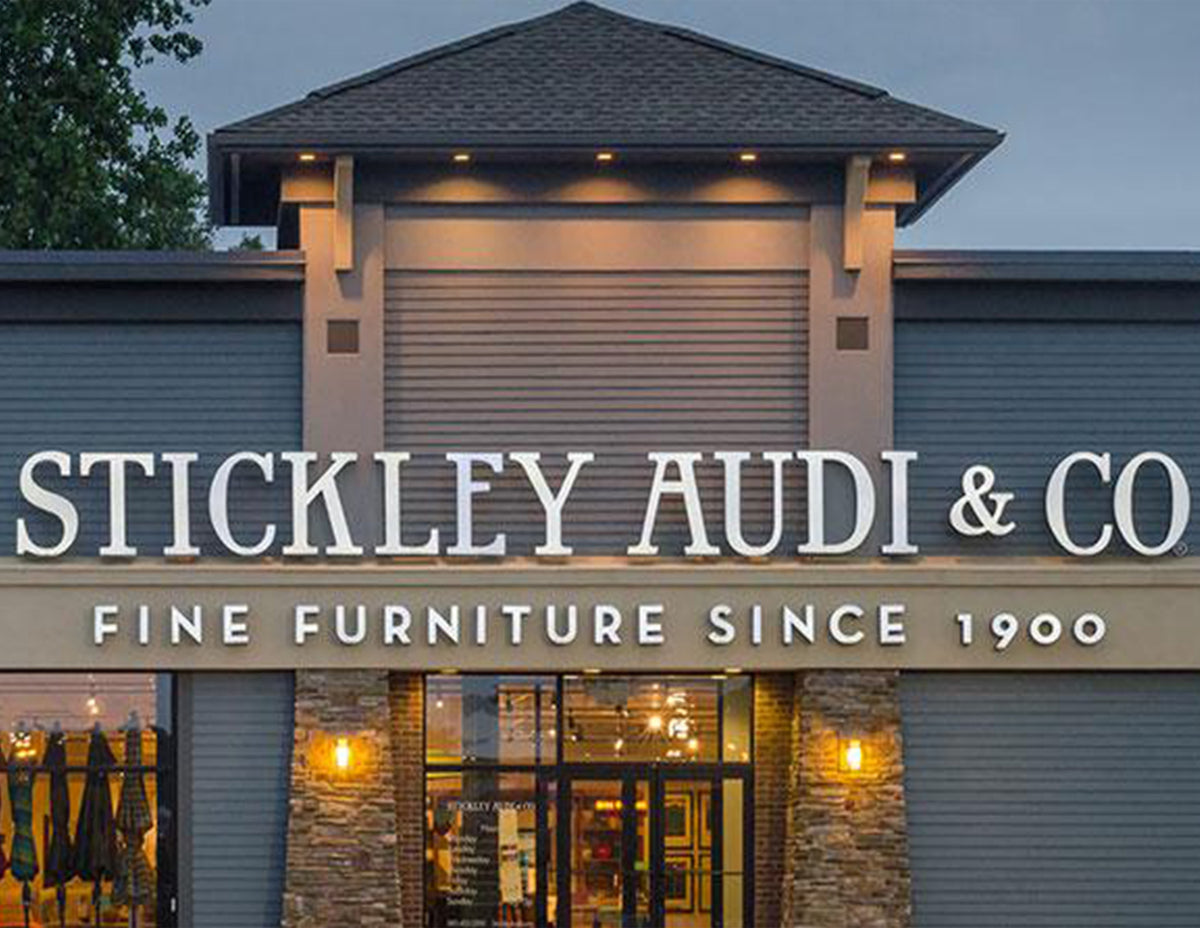 Exterior view of the Stickley Audi & Co. showroom in Rochester, NY, featuring illuminated signage and stone-accented entrance—representing over a century of fine furniture craftsmanship and design excellence since 1900.