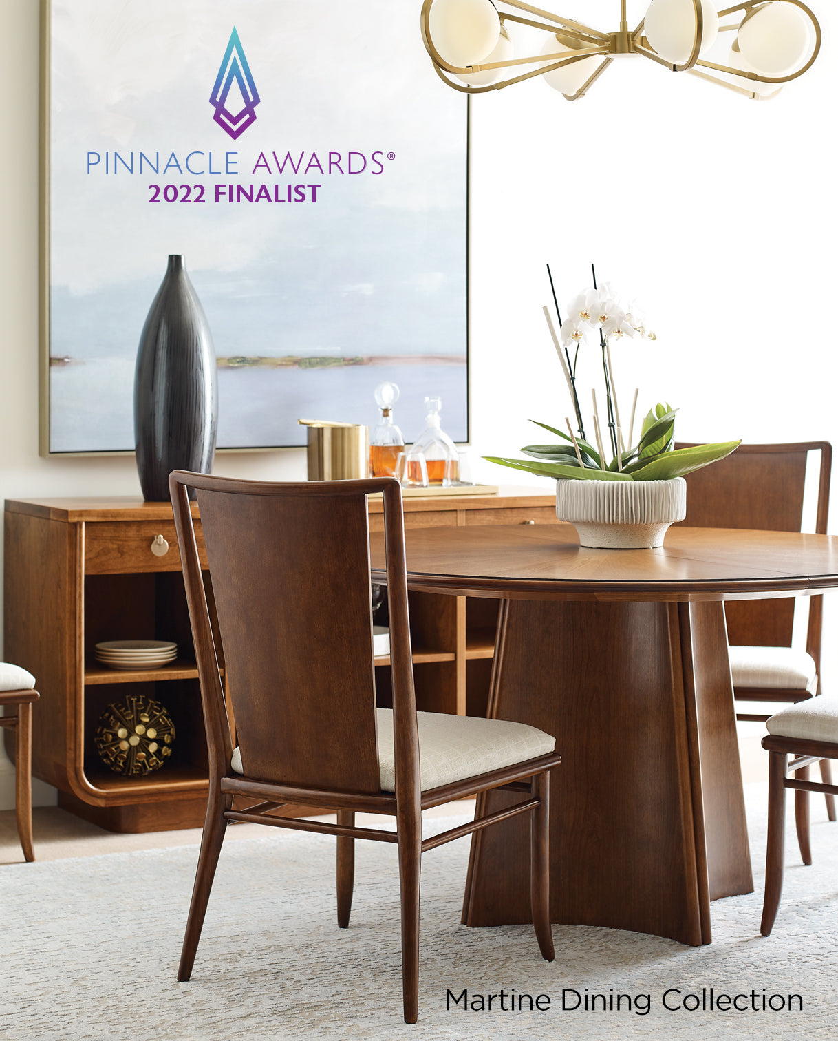 Modern dining room featuring the Pinnacle Awards® 2022 Finalist Martine Dining Collection, with a round pedestal dining table and curved-back wooden chairs upholstered in a light fabric. A contemporary sideboard and oversized abstract artwork complete the space, along with a gold chandelier and a centerpiece of white orchids.