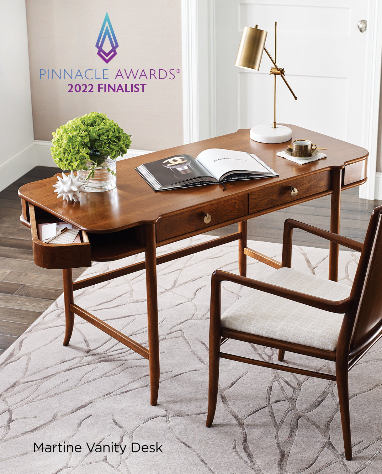 Pinnacle Awards® 2022 Finalist Martine Vanity Desk shown in a bright, modern room. The elegant wooden desk features sculpted edges, a pull-out side tray, and two central drawers, paired with a matching upholstered chair. A gold desk lamp, cup of coffee, open book, and floral arrangement complete the serene workspace.