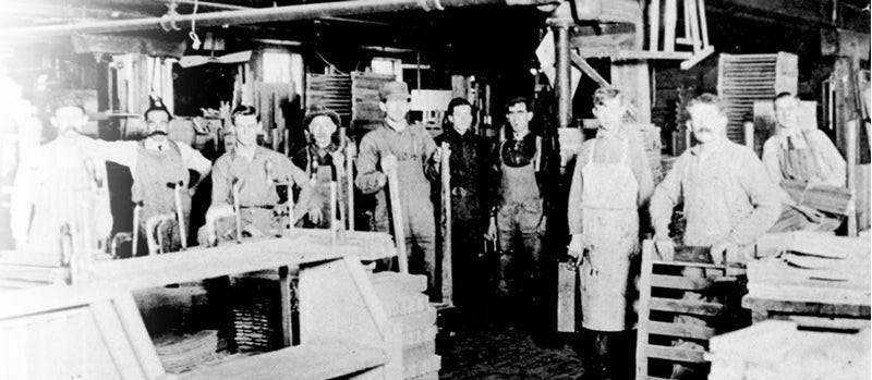 Black-and-white photo of early 20th-century Stickley factory workers standing in a woodshop surrounded by tools, stacks of lumber, and unfinished furniture pieces.