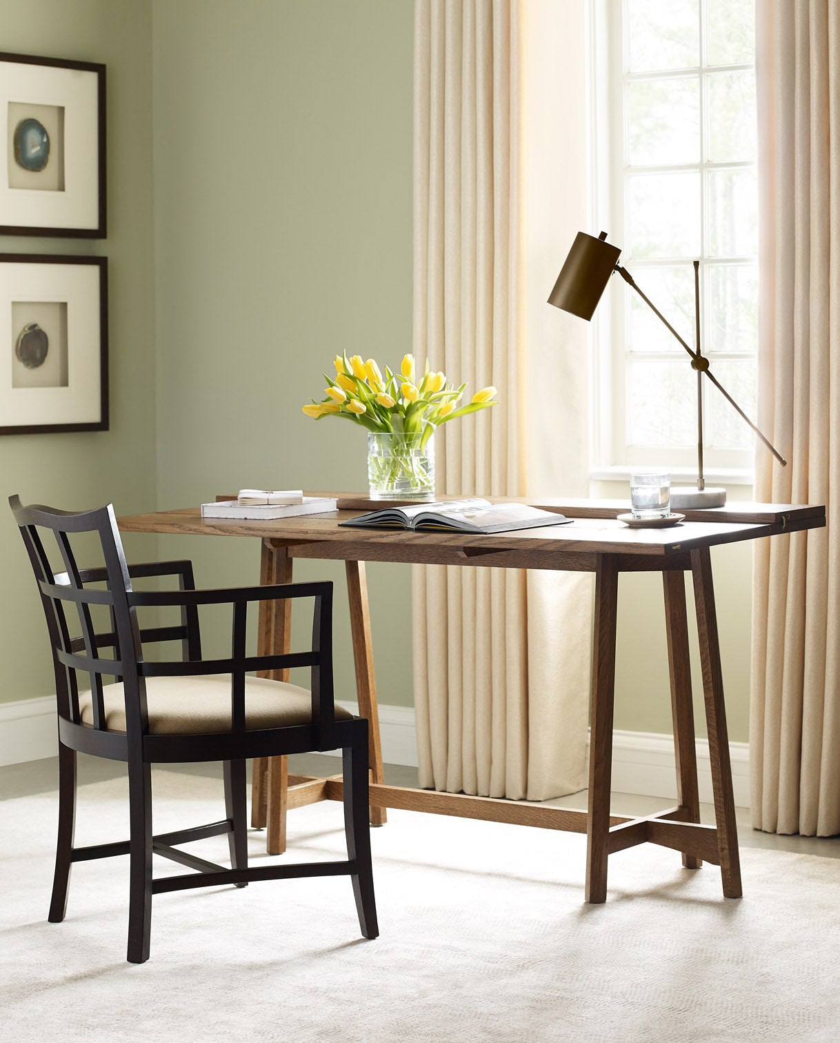 A Surrey Hills Flip-Top Console Table with a Surrey Hills Arm chair slightly pulled out from it. They are in a light green colored room that has a large window with cream colored curtains