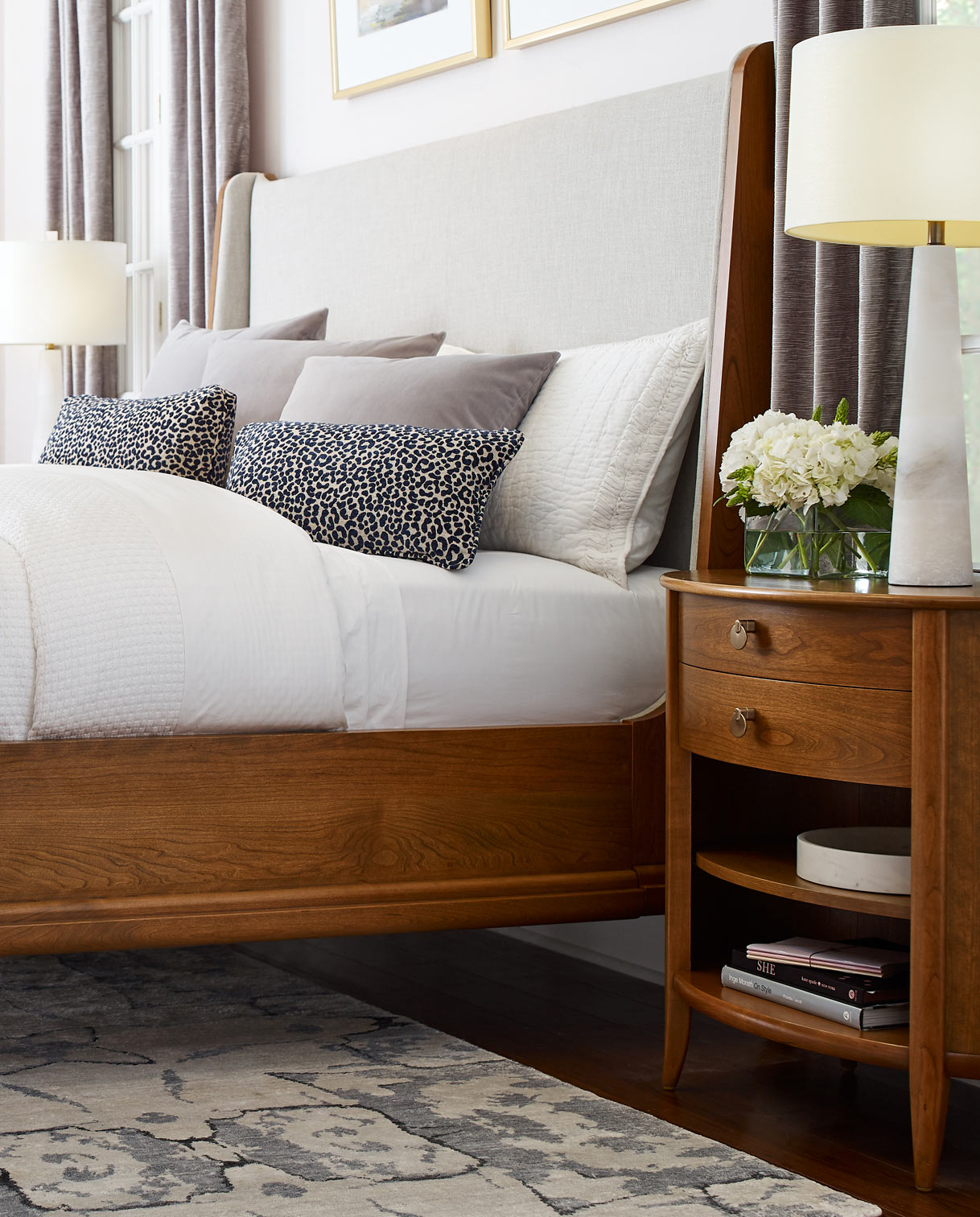Martine nighstand with a white lamp and small vase of white flowers on top, next to a Martine light gray upholstered bed that has a white comforter, black and white decorative pillows, and gray and white pillows