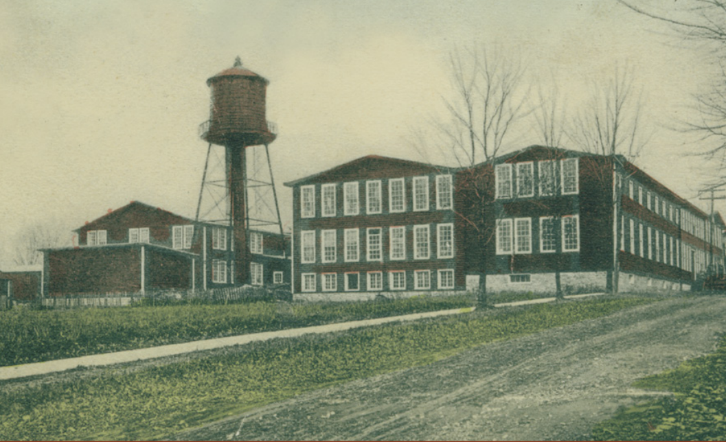 Historic photograph of the original L. & J.G. Stickley factory, showing a large red brick industrial building with rows of tall windows and a prominent water tower on metal supports. The factory sits along a dirt road with bare trees and grassy areas in the foreground, evoking an early 20th-century setting.